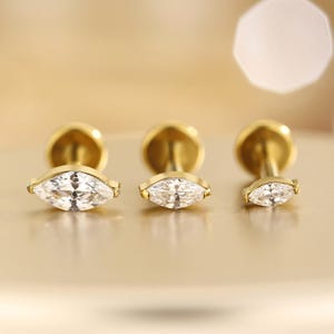 May include: Three gold-tone flat back stud earrings arranged in a row, each featuring a marquise-cut clear white gemstone. The studs vary in size from largest on the left to smallest on the right, with a metallic gold post and circular backing.