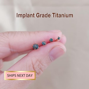 Könnte beinhalten: Vier kleine, goldfarbene Ohrstecker mit dunkelblau-grünen Edelsteinen werden gezeigt. Der Text "Implant Grade Titanium" steht über den Ohrringen. Der Text "SHIPS NEXT DAY" befindet sich in einer rosa Box darunter.