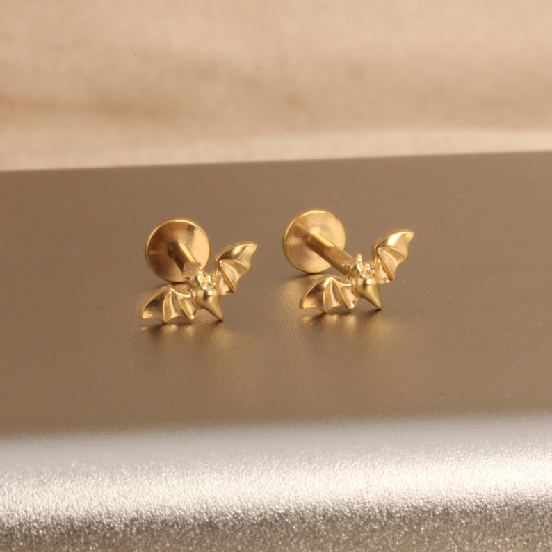 May include: Two gold-colored bat-shaped earrings. Each earring features a small, detailed bat design with outstretched wings and a pointed head. The earrings have a flat, round backing for secure wear.