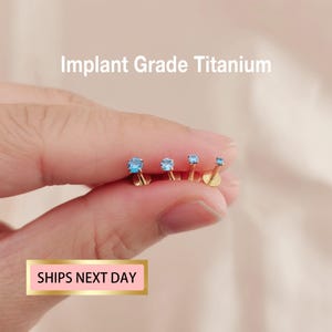 May include: Four gold-coloured, implant-grade titanium earrings with light blue gemstones. The earrings are in various sizes and are held by a person. The text "Implant Grade Titanium" is visible. The text "SHIPS NEXT DAY" is also visible.