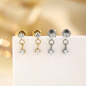 May include: Close-up of three pairs of earrings. Two pairs are gold-toned, and one pair is silver-toned. Each earring features a round, clear gemstone at the top and a dangling square gemstone at the bottom, connected by a small chain.