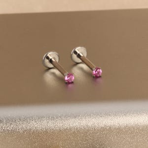 Może przedstawiać: Dwa srebrne kolczyki labret z r&oacute;żowymi kamieniami szlachetnymi. Każdy kolczyk ma płaską, okrągłą podstawę, smukły pręt i mały, okrągły, r&oacute;żowy kamień szlachetny. Biżuteria jest prezentowana na jasnobrązowej powierzchni.