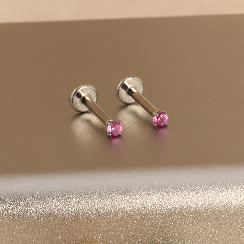 Może przedstawiać: Dwa srebrne kolczyki labret z r&oacute;żowymi kamieniami szlachetnymi. Każdy kolczyk ma płaską, okrągłą podstawę, smukły pręt i mały, okrągły, r&oacute;żowy kamień szlachetny. Biżuteria jest prezentowana na jasnobrązowej powierzchni.