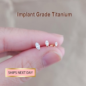 May include: Three gold earrings, each with a clear, marquise-shaped gemstone. The earrings are shown against a blurred backdrop. The text "Implant Grade Titanium" is visible at the top.