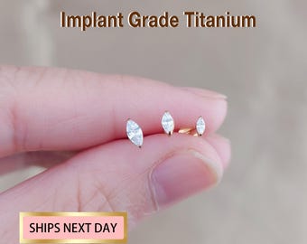 May include: Three gold-toned stud earrings featuring marquise-cut clear stones. The text Implant Grade Titanium is visible at the top. These earrings have a metallic finish and pointed stone shapes, designed for ear piercings with a secure backing.