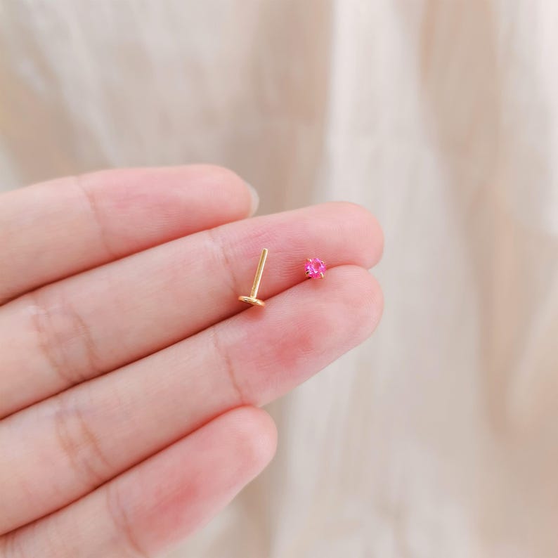 K&ouml;nnte beinhalten: Ein goldfarbener Ohrstecker mit geradem Steg und flachem R&uuml;cken, neben einem kleinen, herzf&ouml;rmigen, leuchtend rosa Edelstein-Ohrstecker. Die Ohrringe werden auf einer hellrosa Oberfl&auml;che pr&auml;sentiert.