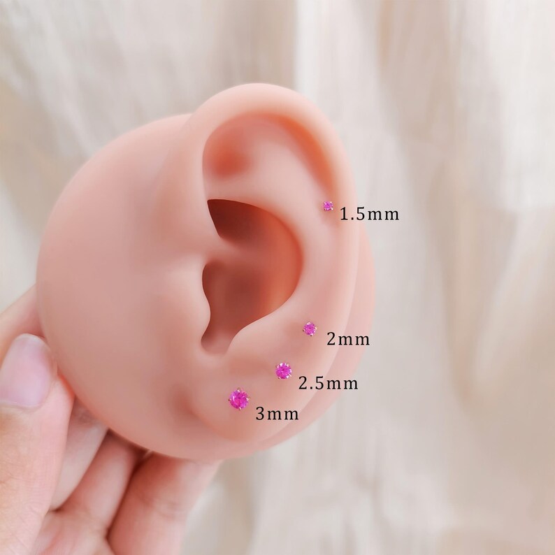 K&ouml;nnte beinhalten: Nahaufnahme eines rosa Ohrmodells mit vier kleinen, runden, rosa Ohrsteckern. Die Ohrringe sind mit ihren Gr&ouml;&szlig;en beschriftet: 1,5 mm, 2 mm, 2,5 mm und 3 mm. Das Ohrmodell wird von einer Hand gehalten.