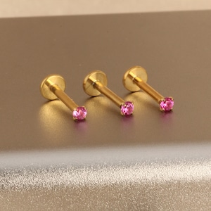 Może przedstawiać: Trzy złote kolczyki do ust lub uszu z małym, okrągłym, r&oacute;żowym kamieniem szlachetnym na końcu każdego pręcika. Kolczyki mają płaskie, okrągłe oparcie. Tło jest w neutralnym, metalicznym kolorze.