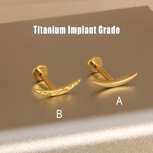May include: Two gold-coloured, crescent-shaped body jewellery pieces. One piece is smooth, and the other has a decorative pattern. The text "Titanium Implant Grade" is above the jewellery. The pieces are labelled "A" and "B".