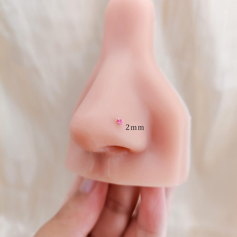 K&ouml;nnte beinhalten: Ein pinkfarbener quadratischer Nasenpiercing, ca. 2 mm gro&szlig;, wird auf einer Schaufensternase gezeigt. Der Stecker hat eine leuchtend pinke Farbe und ist in einem einfachen, minimalistischen Design gehalten. Der Text "2mm" steht neben dem Stecker.