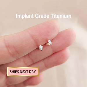 May include: A pair of gold-colored stud earrings, each featuring a marquise-cut, clear gemstone. The earrings are displayed against a light pink background. Text at the top reads "Implant Grade Titanium". A pink rectangle at the bottom says "SHIPS NEXT DAY".