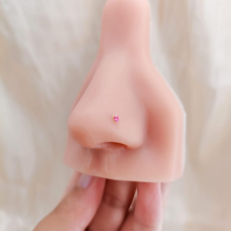 K&ouml;nnte beinhalten: Nahaufnahme einer Nase mit einem kleinen, quadratischen, rosa Edelstein-Nasenpiercing. Der Stecker ist in einem goldfarbenen Metall gefasst. Die Nase hat einen hellen Hautton, der Hintergrund ist neutral.