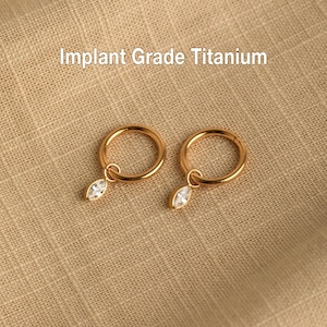 Peut inclure: Deux boucles d'oreilles dorées en forme de cerceau, chacune ornée d'un petit pendentif en forme de marquise transparent. Les boucles d'oreilles sont en titane de qualité implantable. Le texte "Implant Grade Titanium" est visible.