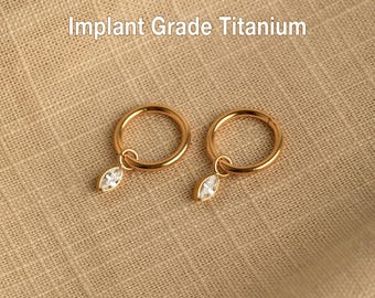 May include: A pair of gold-colored hoop earrings with a small, clear, marquise-cut gemstone charm. The earrings are made of implant grade titanium. The text "Implant Grade Titanium" is visible in the image.
