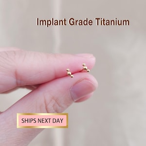 May include: A pair of small, gold-colored stud earrings. Each earring features three small, round beads arranged in a cluster. The text "Implant Grade Titanium" is displayed above the earrings.