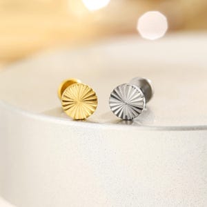 Könnte beinhalten: Nahaufnahme von zwei Ohrsteckern. Einer ist goldfarben, der andere silberfarben. Beide Ohrringe haben ein kreisförmiges Design mit Sonnenstrahlen. Sie liegen auf einer weißen Oberfläche mit unscharfem Hintergrund.