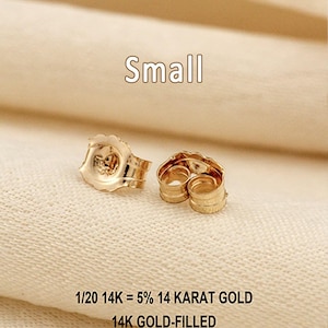 Puede incluir: Dos cierres de pendiente peque&ntilde;os de color dorado con un dise&ntilde;o de borde festoneado. El texto Small, 1/20 14K = 5% 14 KARAT GOLD y 14K GOLD-FILLED es visible, indicando la composici&oacute;n del material de estos componentes de joyer&iacute;a.