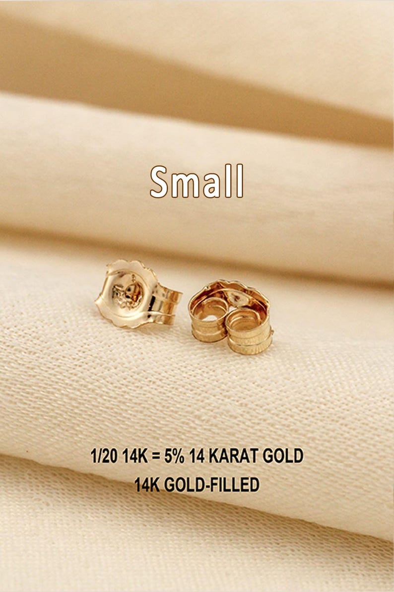 Puede incluir: Dos cierres de pendiente peque&ntilde;os de color dorado con un dise&ntilde;o de borde festoneado. El texto Small, 1/20 14K = 5% 14 KARAT GOLD y 14K GOLD-FILLED es visible, indicando la composici&oacute;n del material de estos componentes de joyer&iacute;a.