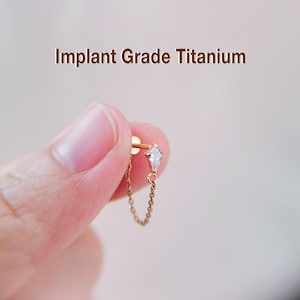 Peut inclure: Une boucle d'oreille dorée avec une petite gemme transparente en forme de diamant et une chaîne délicate. La boucle d'oreille est maintenue par une tige. Le texte "Implant Grade Titanium" est affiché au-dessus.