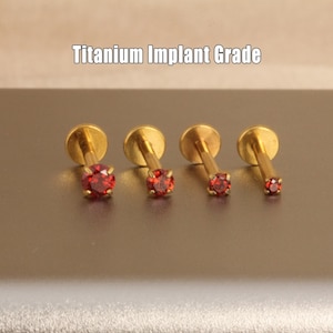 May include: Four gold-coloured, titanium implant grade lip piercing studs with red gemstones. The studs are arranged in a row, with the gemstones decreasing in size. The text "Titanium Implant Grade" is above the studs.