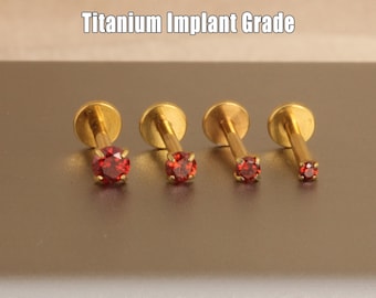 May include: Four gold-coloured, titanium implant grade lip piercing studs with red gemstones. The studs are arranged in a row, with the gemstones decreasing in size. The text "Titanium Implant Grade" is above the studs.