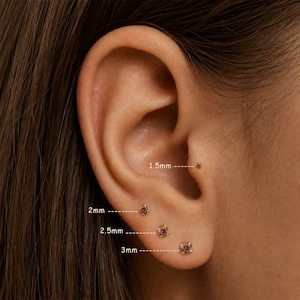 May include: Close-up of an ear with four small, gold-colored stud earrings. The earrings are adorned with small, round, reddish-brown gemstones. The sizes are labeled as 1.5mm, 2mm, 2.5mm, and 3mm.