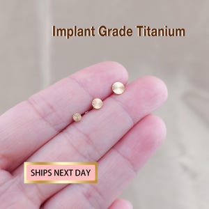 May include: Three gold-colored, circular stud earrings with a textured, radial design are displayed. The earrings are of varying sizes, with the largest at the top. The text "Implant Grade Titanium" is above the earrings.