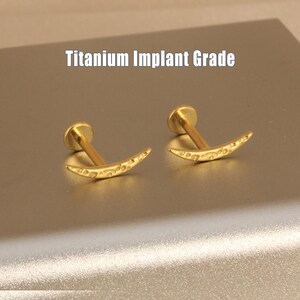 Könnte beinhalten: Zwei goldfarbene, halbmondförmige Ohrringe mit einem dekorativen Muster. Jeder Ohrring hat einen flachen, runden Pfosten. Der Text "Titanium Implant Grade" ist in Weiß zu sehen.