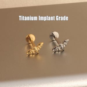 May include: Two titanium implant grade labret studs. One is gold-colored, the other is silver-colored. Each stud features a bat-shaped charm. The text "Titanium Implant Grade" is displayed above the studs.