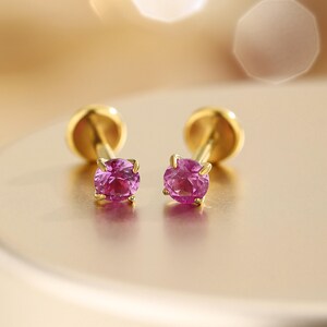 K&ouml;nnte beinhalten: Ein Paar goldene Ohrstecker, jeweils mit einem leuchtend rosa Edelstein. Die Ohrringe haben ein klassisches Design mit einem runden, polierten Goldr&uuml;cken und einer Vier-Zacken-Fassung, die den rosa Stein h&auml;lt. Die Edelsteine sind tiefrosa.
