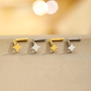 May include: Four nose studs with star-shaped designs. Two are gold-colored, and two are silver-colored. The studs are arranged in a row, with the star-shaped portion facing forward. The background is a neutral, warm tone.