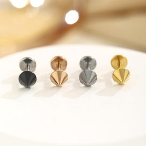 May include: A collection of six stud earrings in various metallic finishes. The earrings feature a round post with a cone-shaped front. Colors include black, gold, silver, and rose gold. The earrings are displayed on a white surface.