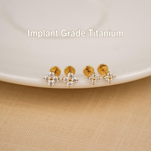 Peut inclure: Quatre boucles d'oreilles à clous dorées en forme de fleur, ornées de pierres scintillantes et transparentes. Elles sont présentées sur une assiette blanche. Le texte "Implant Grade Titanium" est au-dessus.