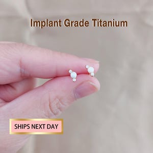 Könnte beinhalten: Ein Paar kleine, goldfarbene Ohrringe mit drei weißen, runden Steinen. Die Ohrringe werden in einer Hand gehalten. Der Text "Implant Grade Titanium" befindet sich darüber.