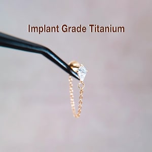 Peut inclure: Une boucle d'oreille dorée avec une pierre précieuse transparente en forme de losange et une chaîne délicate. La boucle d'oreille est tenue par une pince à épiler, avec le texte "Implant Grade Titanium" au-dessus.