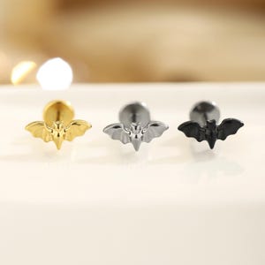 Könnte beinhalten: Drei fledermausförmige Ohrstecker in Gold, Silber und Schwarz. Jeder Ohrring hat ein detailliertes Fledermausdesign mit ausgebreiteten Flügeln. Die Ohrringe haben eine flache Rückseite und einen Stecker zum Durchstechen.