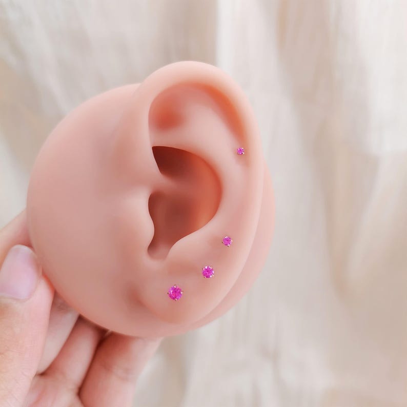 K&ouml;nnte beinhalten: Nahaufnahme eines rosa Ohrmodells mit vier kleinen, runden Ohrsteckern aus rosa Edelsteinen. Die Ohrringe sind in einer Reihe am Ohr angeordnet. Das Ohrmodell wird von einer Hand gehalten, der Hintergrund ist ein weicher, cremefarbener Stoff.
