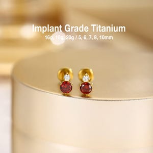 May include: A pair of gold-tone stud earrings featuring a round red gemstone topped with a smaller clear white stone. The text Implant Grade Titanium 16g, 18g, 20g / 5, 6, 7, 8, 10mm is displayed above the earrings, indicating material and size options.