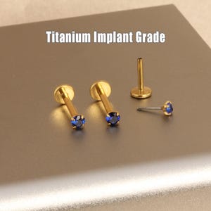 May include: Gold-colored titanium implant grade lip piercing studs with blue gemstones. The image shows several pieces, including two studs, a post, and a single gemstone stud. The text "Titanium Implant Grade" is displayed at the top.