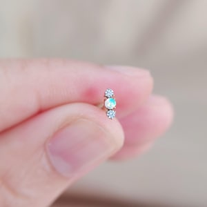 May include: A small, silver-toned earring with three decorative stones. The center stone is an opal, flanked by two clear, sparkling stones. The earring is held between fingers against a blurred background.