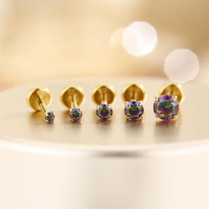 May include: A collection of gold-colored stud earrings with iridescent, round gemstones. The earrings are arranged in a row, showcasing varying sizes of the gemstones. The background is a soft, blurred golden hue.