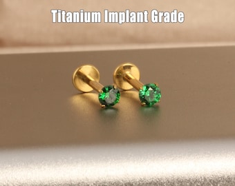 May include: Two gold-colored, stud earrings with a small, round, green gemstone. The earrings have a flat disc on one end and a post with a green gem on the other. The text "Titanium Implant Grade" is above the earrings.