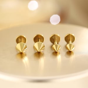 May include: Four gold-colored, cone-shaped stud earrings. The earrings have a flat, round backing and a pointed, geometric design on the front. The earrings are displayed on a gold-toned surface.