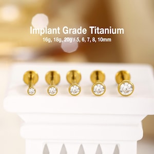 Könnte beinhalten: Fünf goldfarbene Labret-Stecker mit klaren, runden Edelsteinen in verschiedenen Größen. Der Text lautet Implant Grade Titanium 16g, 18g, 20g / 5, 6, 7, 8, 10mm. Jeder Stecker hat eine flache Rückseite und einen eingefassten Stein.