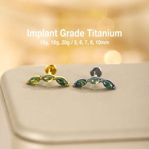 May include: Two curved barbell earrings made of implant grade titanium, one gold-toned and one silver-toned. Each features three marquise-cut green stones. Text reads: Implant Grade Titanium, 16g, 18g, 20g / 5, 6, 7, 8, 10mm.