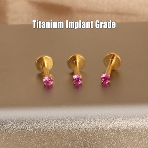 May include: Three gold-coloured, titanium implant grade lip or ear studs, each with a small, round, pink gemstone. The studs have a flat disc on one end and a post with a gemstone on the other. The text "Titanium Implant Grade" is above the studs.