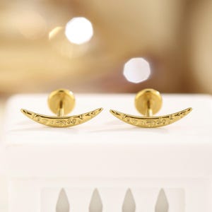 May include: A pair of gold-colored crescent moon earrings. Each earring features a curved, engraved design with a post backing. The earrings are displayed on a white surface with a blurred, warm-toned background.