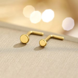 May include: Two gold-colored nose studs with a flat, circular design. The studs are L-shaped, with a small, round disc at the end of the L. The background is a neutral, textured surface with soft, blurred highlights.