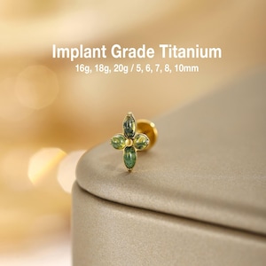 May include: A gold-tone stud earring featuring a four-petaled flower design with green marbled stones. The text Implant Grade Titanium 16g, 18g, 20g / 5, 6, 7, 8, 10mm is displayed above the earring, indicating the material and available sizes.