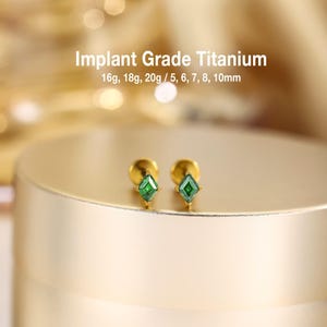 May include: A pair of gold-tone stud earrings featuring kite-shaped green stones. The text Implant Grade Titanium 16g, 18g, 20g / 5, 6, 7, 8, 10mm is visible above the earrings, indicating the material and available gauge and length sizes.